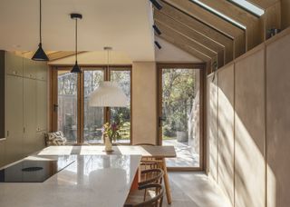 a small kitchen extension with plastered walls and wooden trusses. The kitchen has an slanted extension and in the kitchen is a dining table as well as an island with bar stools.