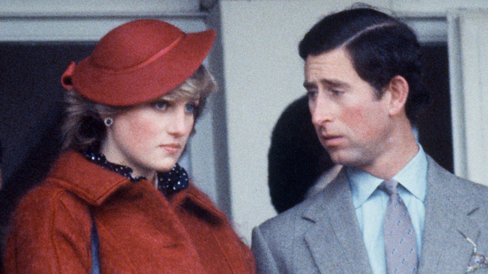 Princess Diana wears a red mohair coat with a matching hat while King Charles wears a light gray suit with a blue shirt and looks at his wife with a worried expression