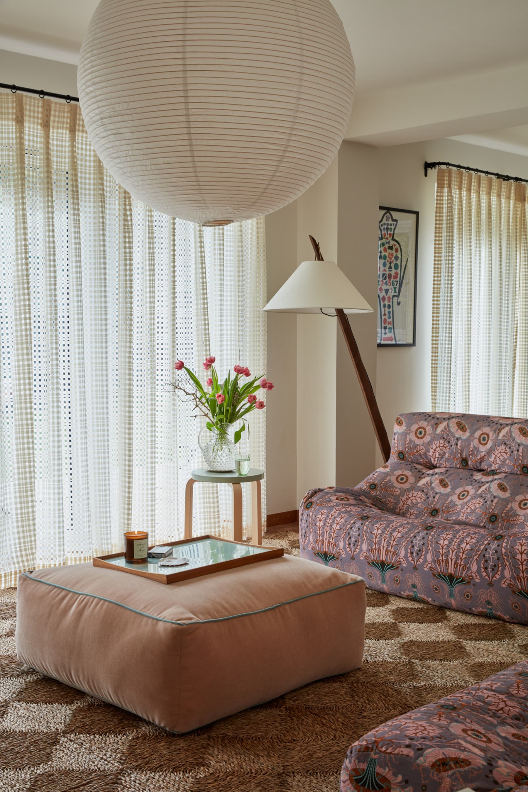 modern living room with perforated curtains, paper lantern, ottoman, checkered jute rug, and pink floral sofa with leaning floor lamp and side table with vase of tulips
