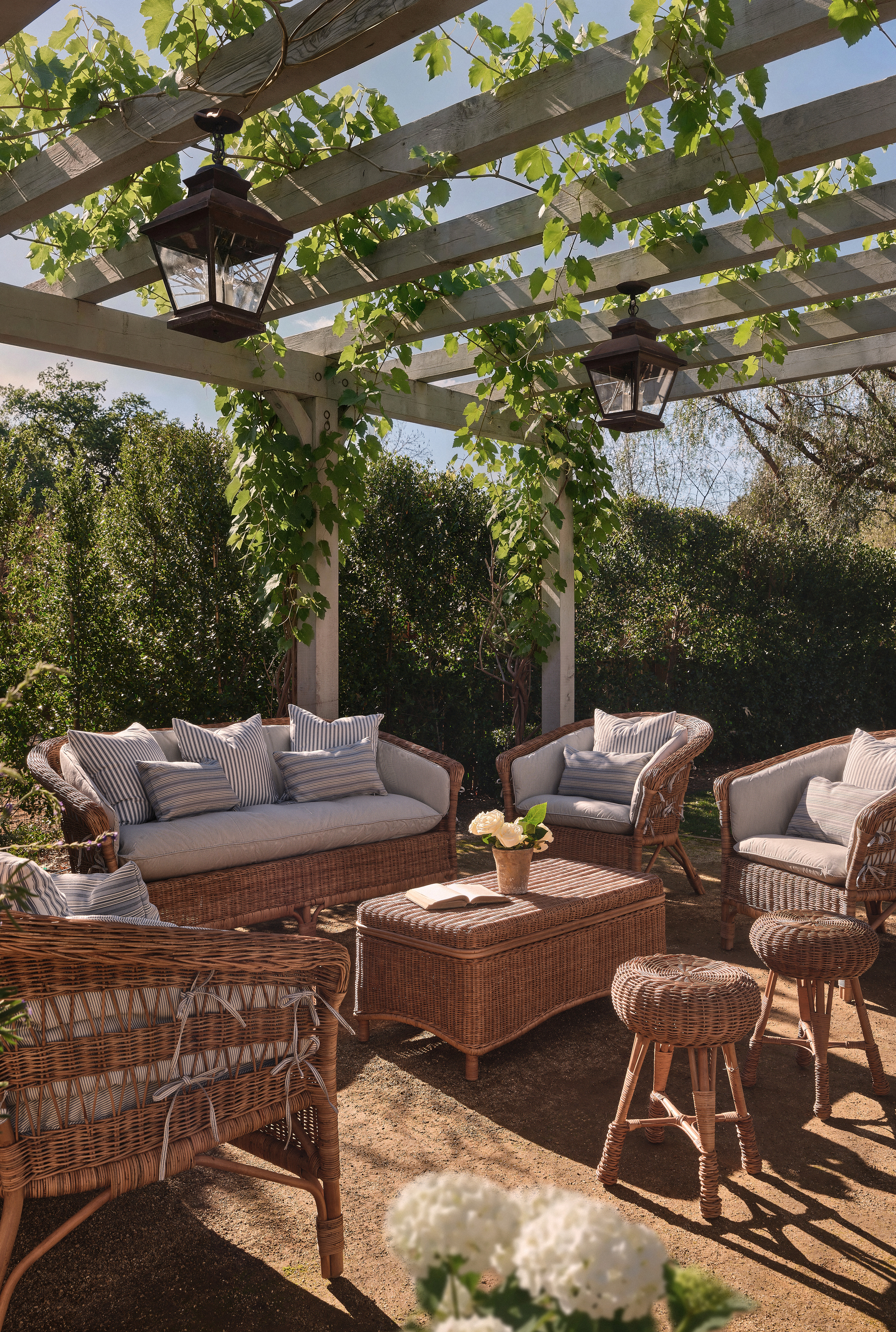 a pergola draped in grapevines with rattan armchairs, sofa, coffee table and stools with blue-and-white striped cushions
