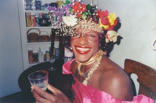 Marsha P. Johnson, wearing a flower crown, sits in a kitchen while smiling and holding a glass, in a still from 'The Death and Life of Marsha P. Johnson'