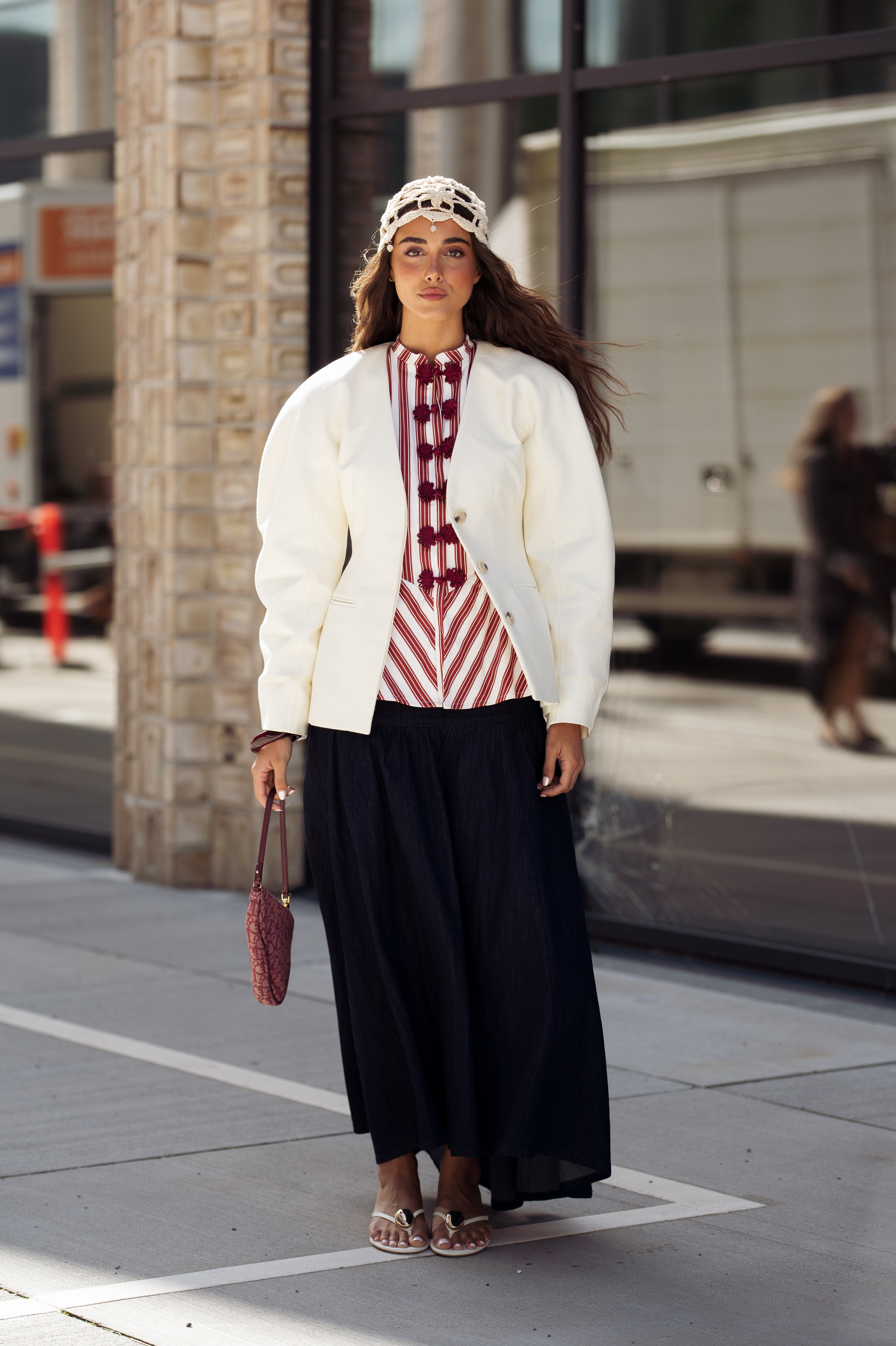 A guest wears long black maxi skirt, red and white top, white jacket and white lace hat outside the Herskind show during Copenhagen Fashion Week