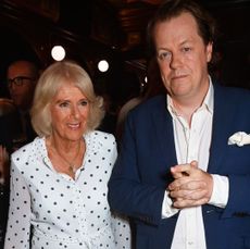 Tom Parker Bowles wearing a blue suit standing next to his mother Queen Camilla who is smiling and wearing a white polkadot dress