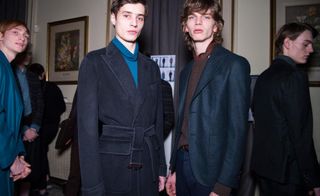 2 male models in dark suit jackets stand in a room full of people