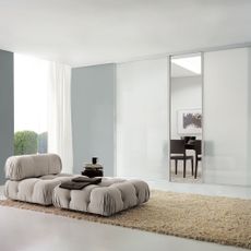 a white living space with full length window, a light coloured buttoned mattress seat on top of a long shaggy cream rug, and a frosted room divider sectioning off a dining area