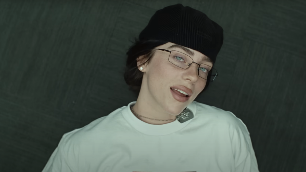 Billie Eilish sitting back in chair with glasses, a cap and white T-shirt in Birds of A Feather music video