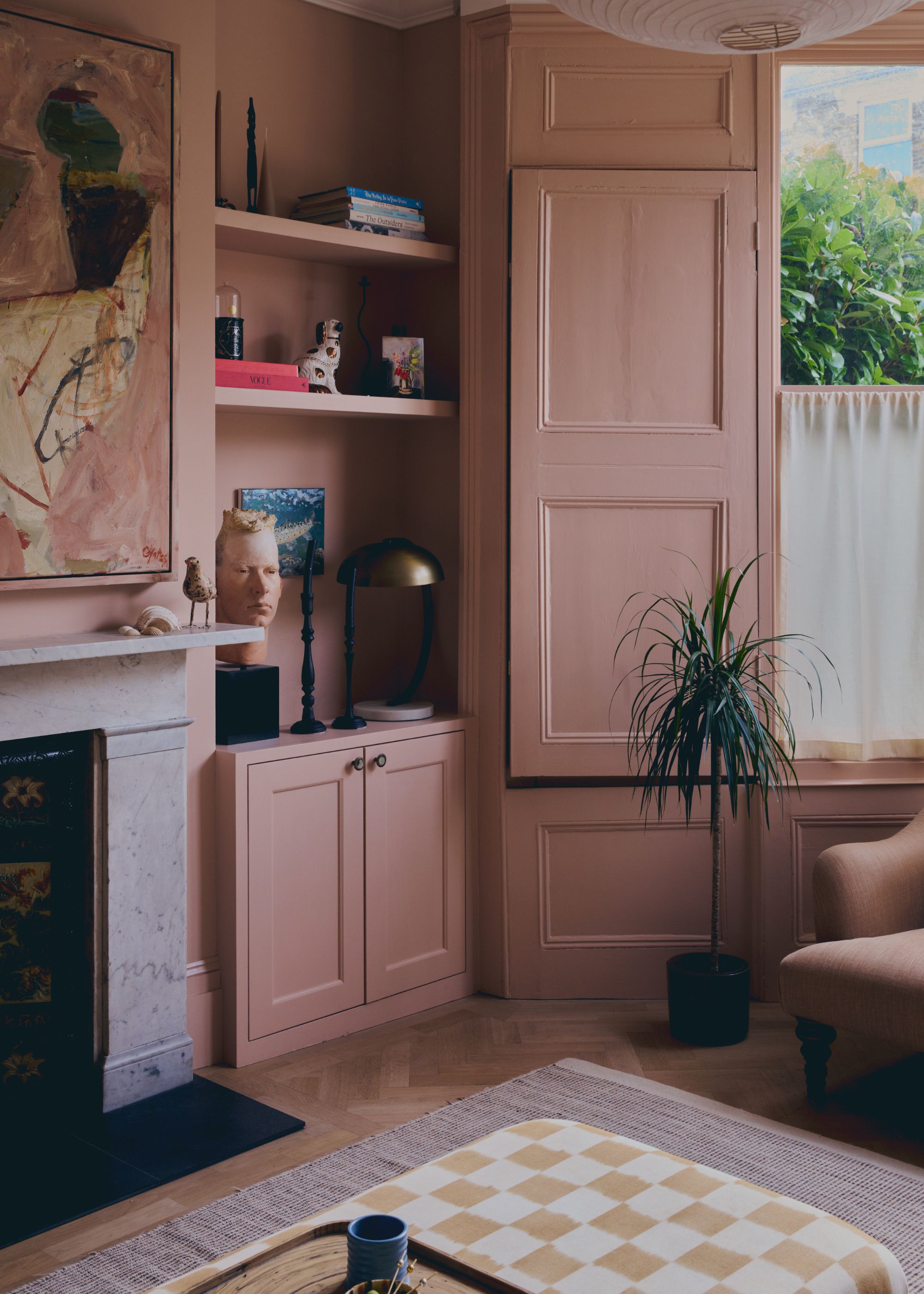 A living room color-drenched plaster pink with a bay window, fireplace, checkered ottoman, and abstract artwork on the wall.