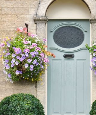 A pale blue door with round mottled glass window, steel gray letterbox and two large hanging baskets on stone walls filled with purple, pink and orange flowers