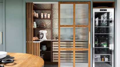 A small walk-in pantry with a sliding wood and fluted glass door in a green kitchen