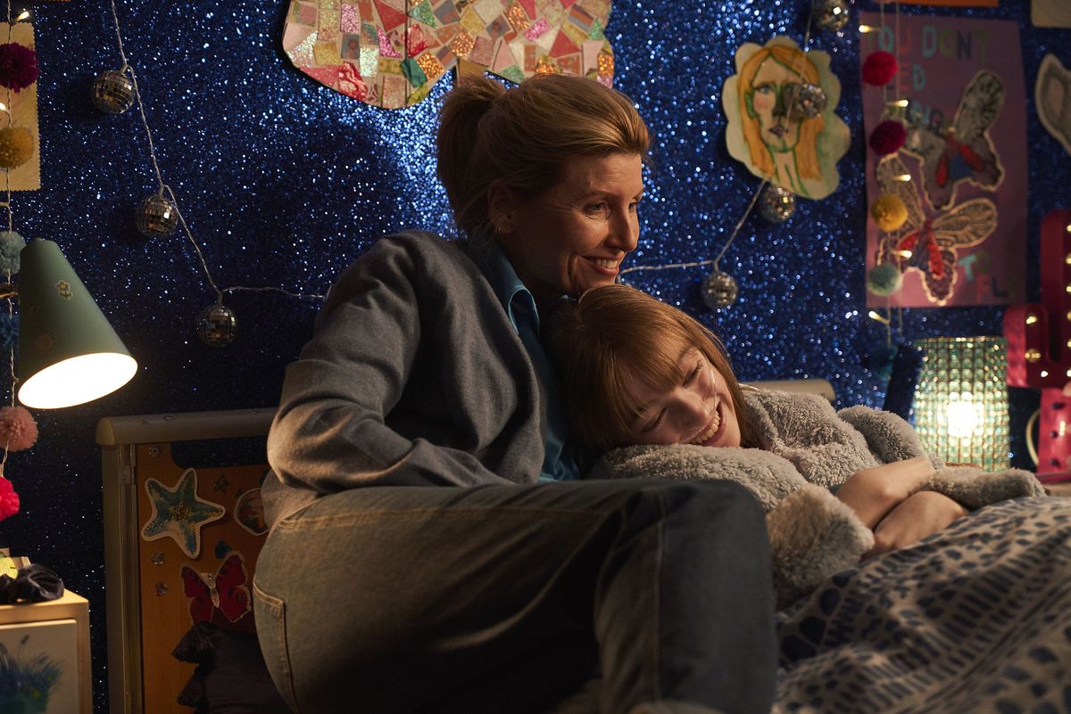 Best Interests cast Sharon Horgan and Niamh Moriarty sitting on a bed together with a blue sparkly background. 