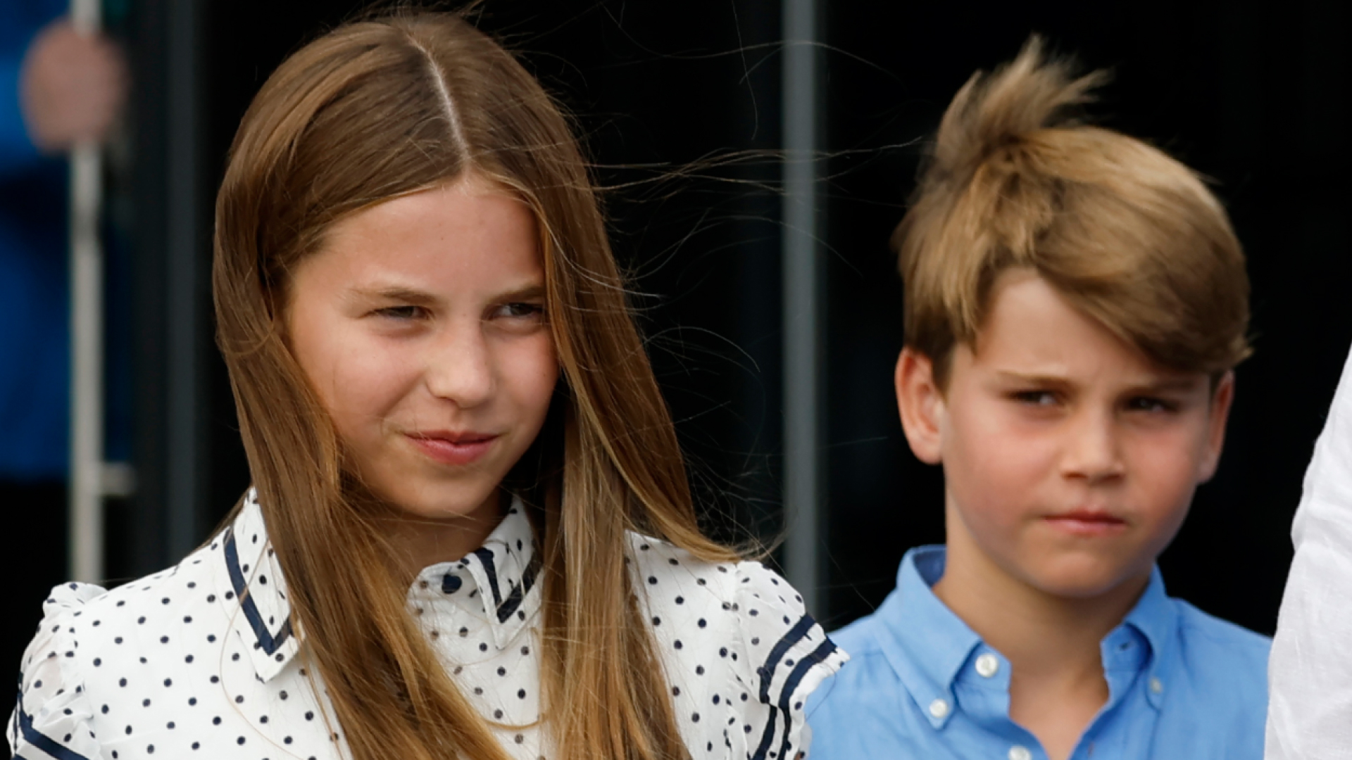 Princess Charlotte has long dark blonde hair and wears a white and black polka dot dress while Princess Louis wears a blue shirt