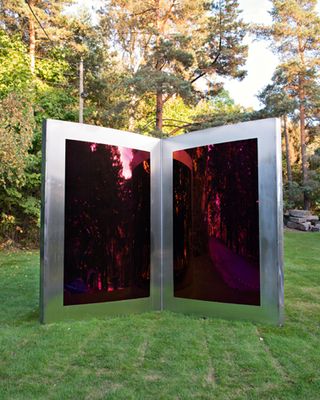 A sculpture in the form of an open book with silver metal edges and red reflective glass insidee. Photograph on green fields durng the day with trees in the background