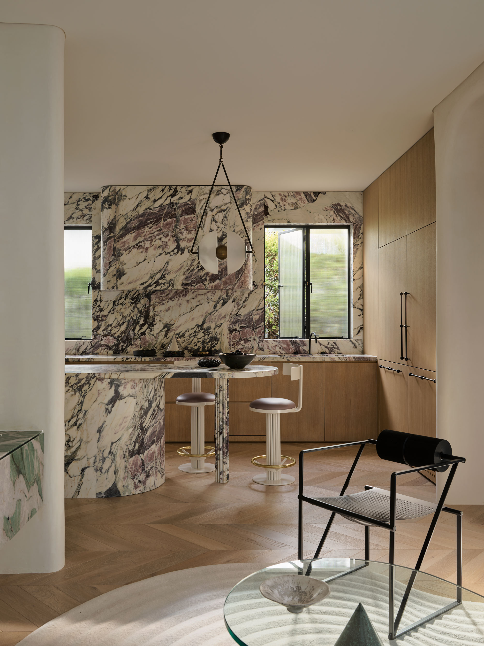 Kitchen space with marble work tops and full marble backsplash on the far wall, light toned wooden cupboards and bar stools.