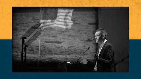A man stands in front of a lectern with a projection of a U.S. flag on the moon appears in the background.