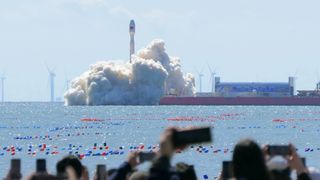 spectators watch a white rocket launch into a blue sky from a ship anchored at sea, not far offshore