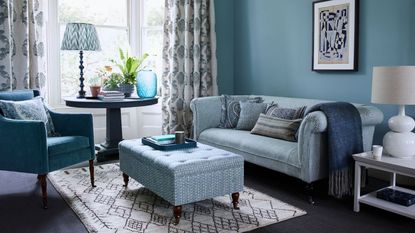 A blue living room with brown wooden floors, and large bay windows with patterned curtains. There's a blue couch, blue upholstered chair and blue patterned storage ottoman, on top of a patterned rug, and a circular table under the window with a lamp, vase and house plant. To the right of the sofa is a dark blue throw, white table and white ceramic lamp with white shade.