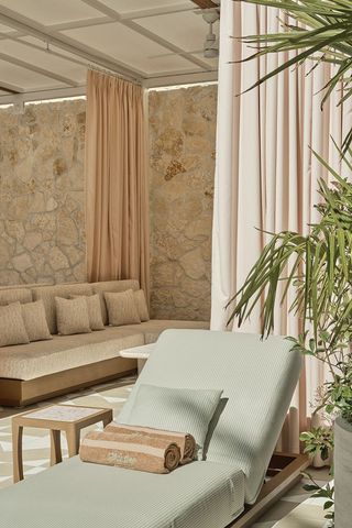 A pool patio of a luxury hotel features earthy shaded upholstered furniture including reclining chairs and banquettes, curtains, and stone walls as well as floors.