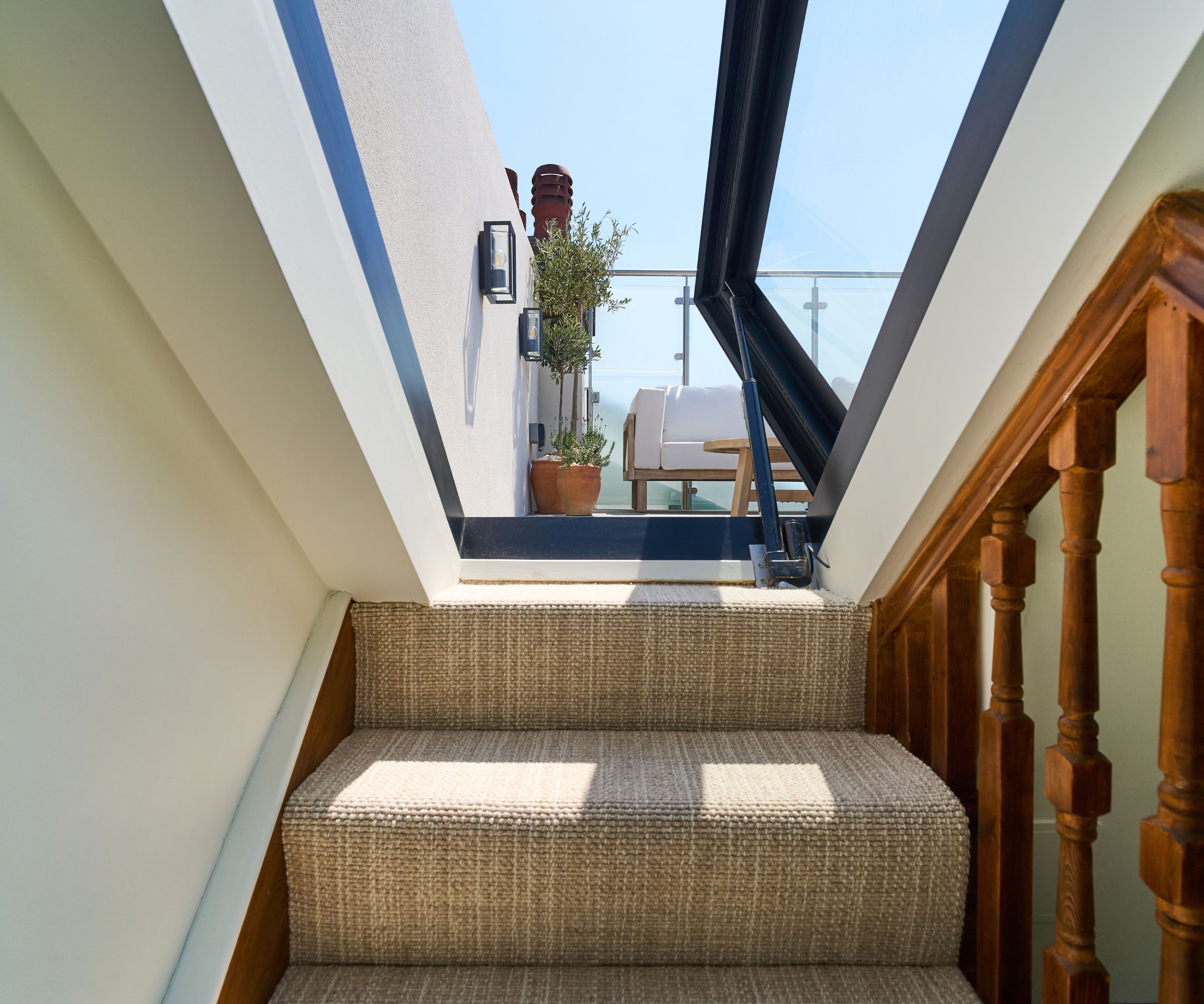 View from staircase looking up through a large glazed hatch to a rooftop terrace with seating and potted plants in sunlight.