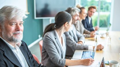 A group of workers meets at a conference table. In the foreground, an older worker looks at the camera knowingly.