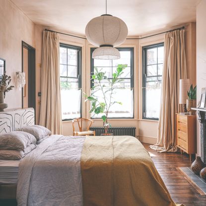 A pink limewash-painted bedroom with a large bay window dressed with tonal full-length pooling curtains
