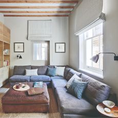 A white TV room with a corner modular sofa in a dark blue linen upholstery with a brown leather ottoman used as a coffee table