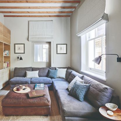 A white TV room with a corner modular sofa in a dark blue linen upholstery with a brown leather ottoman used as a coffee table
