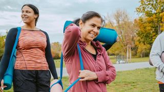 Two women smiling in workout clothes, holding yoga mats walking across grass in the local park