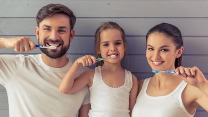 Parents and a small child brush their teeth together.