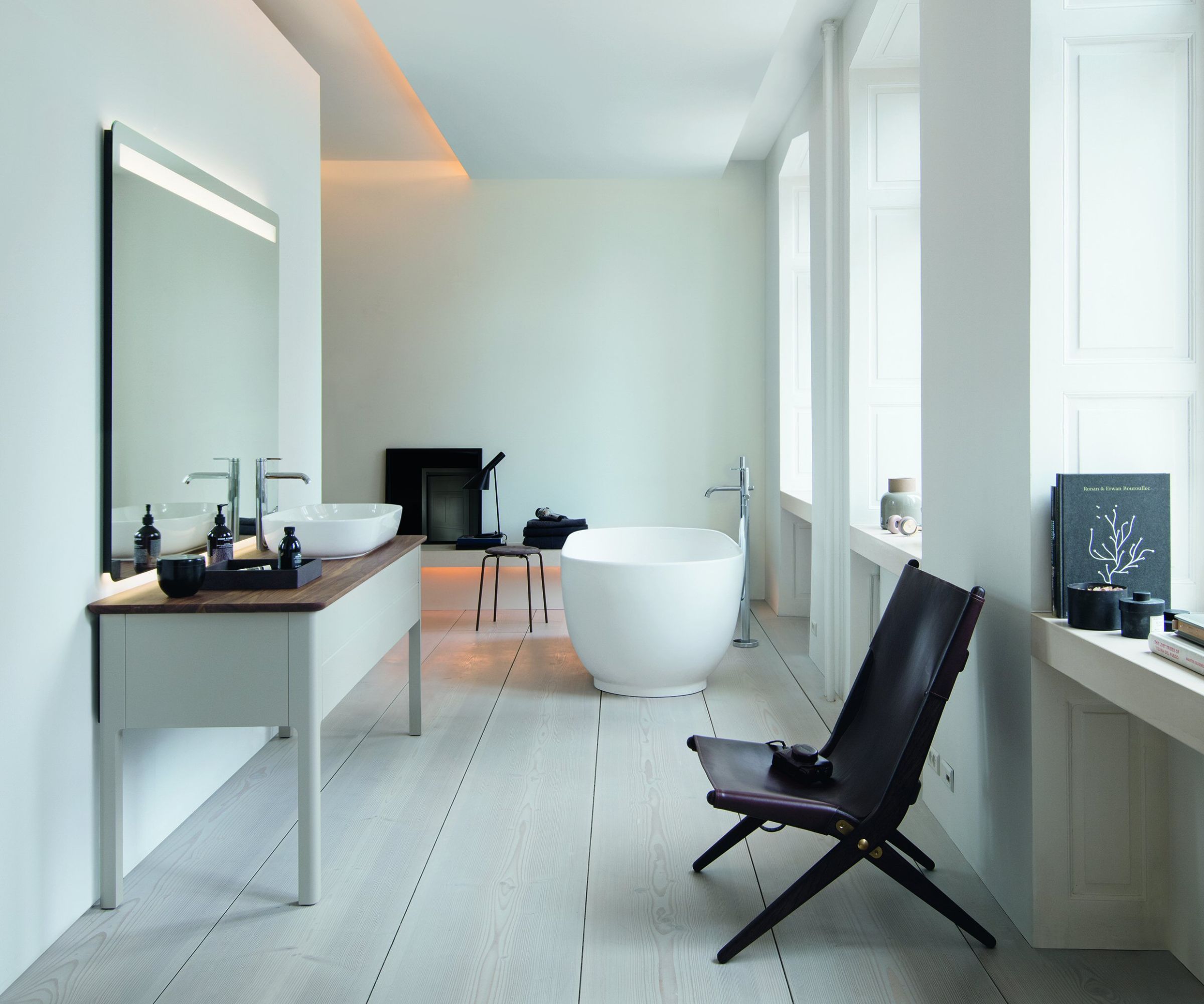 A spacious, minimal Scandinavian bathroom with white walls, wide pale ash floorboards and tall windows. A white console vanity unit with a walnut worktop and twin vessel basins sits beneath a lit mirror, while a sculptural white freestanding bath with a chrome floor-mounted tap occupies the far end.