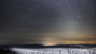 A meteor shower is pictured against a starry sky and picturesque foreground