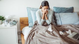 A woman with long dark hair blows her nose in bed because she is sick