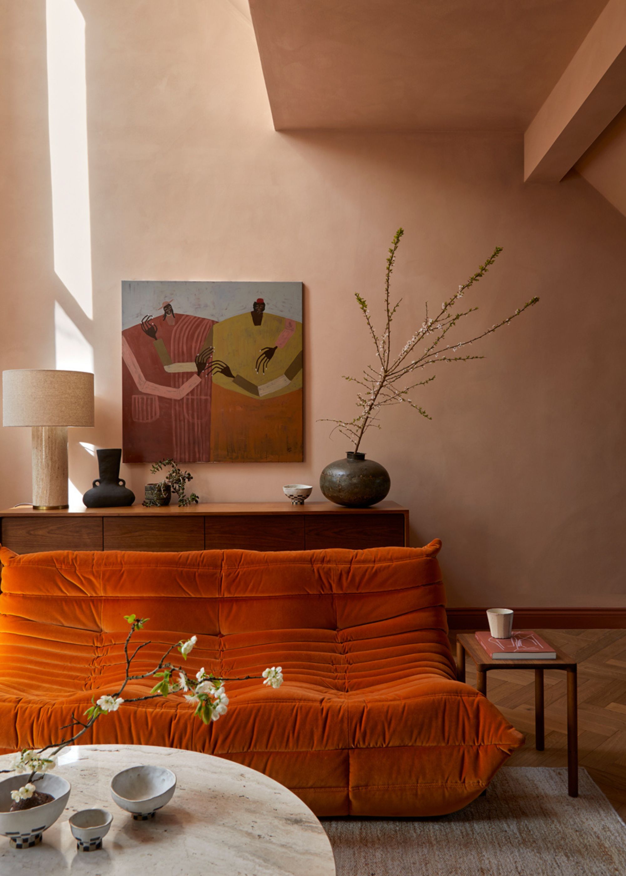 A living room with dusty pink walls, a painting, a sideboard with a vase and a branch, a lamp, a orange Togo, a wood side table, and a table with ikebana