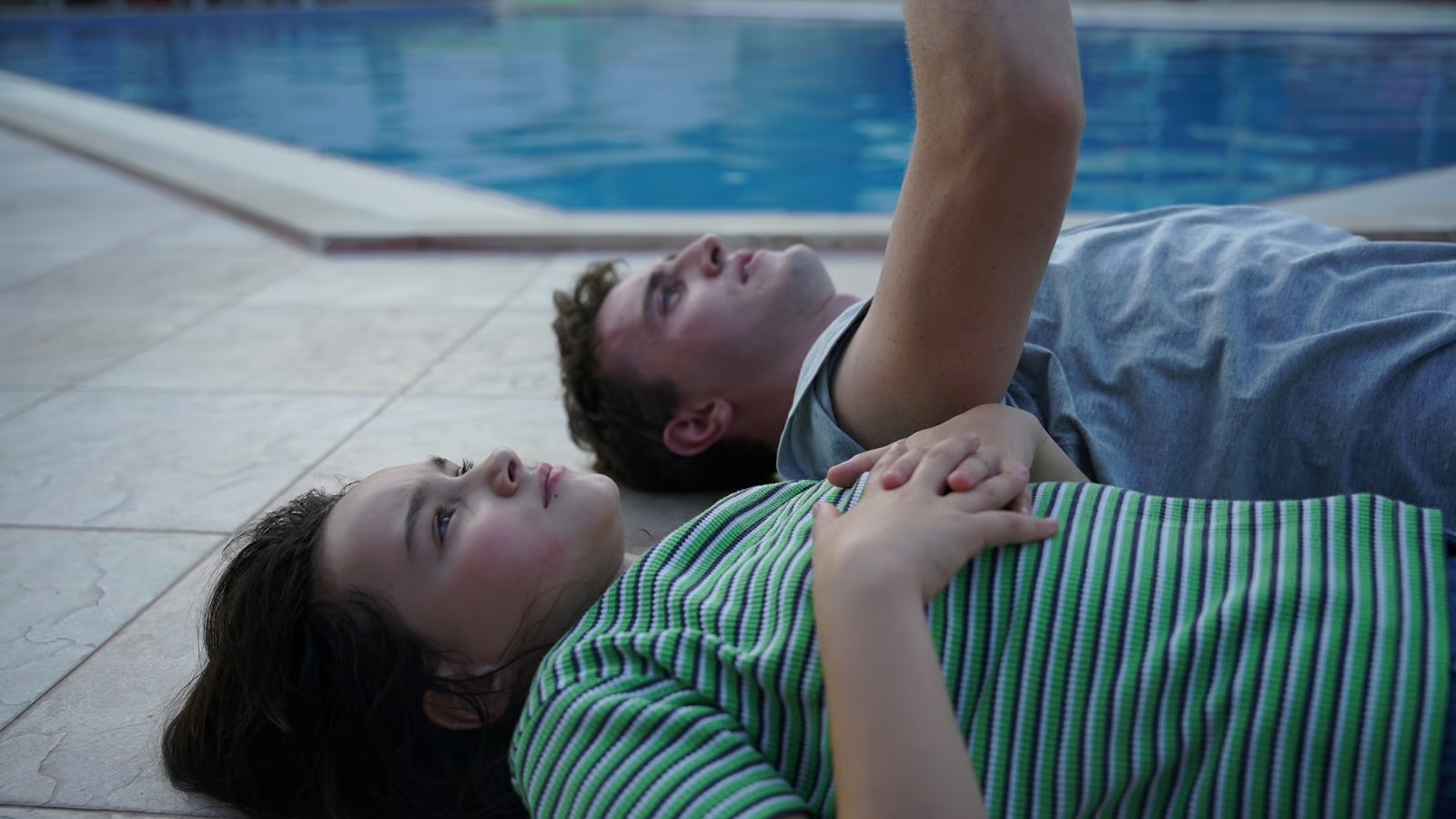 frankie corio and paul mescal lie on their backs outside of a pool looking up at the sky in a still from aftersun
