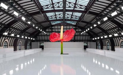 A large red anthurium flower stands in the middle of an all white show space inside a large exhibition room.