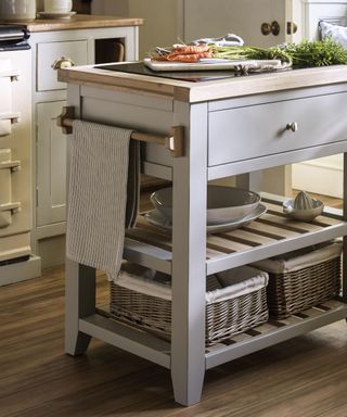 A small gray kitchen island with a wooden surface with carrots on top, a ple with a tea towel on the side of it, and two shelves underneath