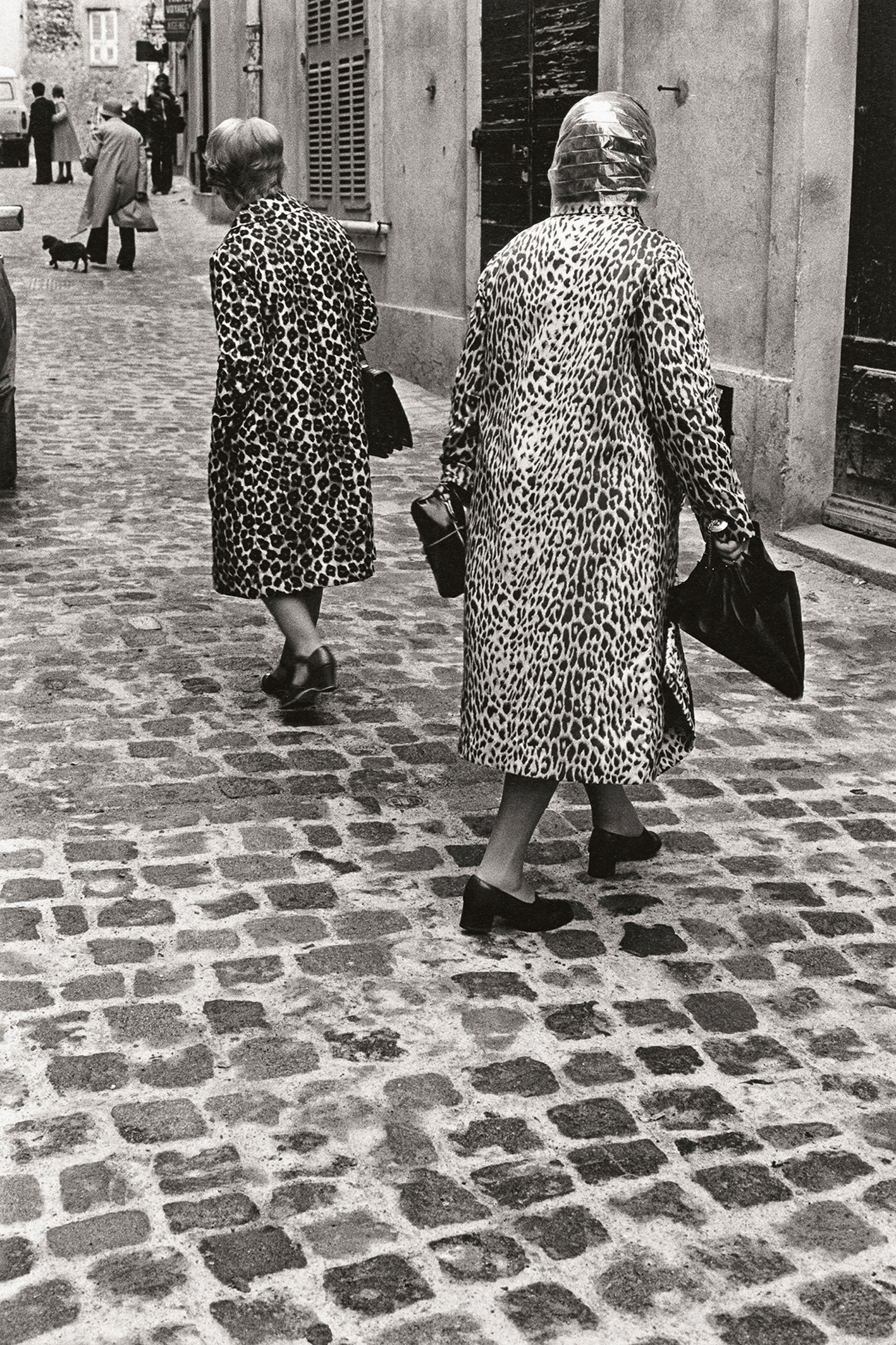 Black and white image of two women in leopard print coats walk down a cobblestone street, shadowed by others in the bustling urban scene