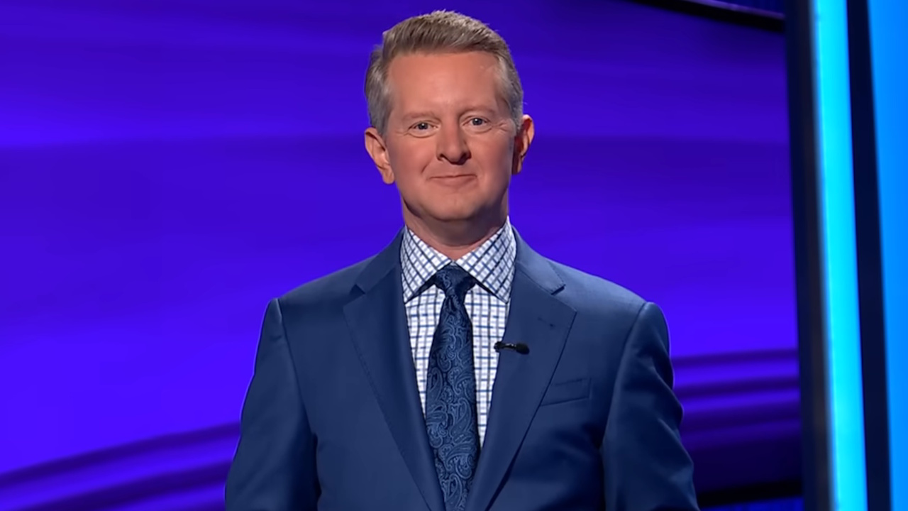 Ken Jennings wears a textured blue paisley tie over a patterned shirt and navy coat on Jeopardy. 