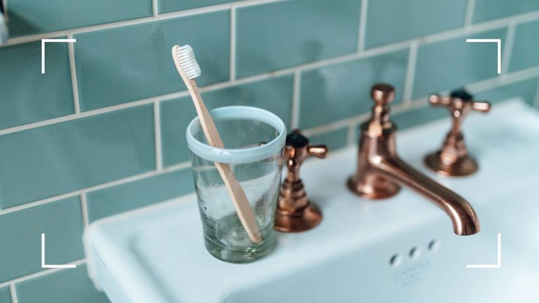 Wooden toothbrush in a glass on the side of a white enamel bathroom sink, unused, with blue and white tiles behind to illustrate how often should you brush your teeth
