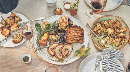 Dinner table with platter of rolled pork in the center and plates of vegetables with a hand holding a glass of red wine