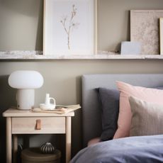IKEA DEJSA table lamp in a calm, neutral toned bedroom, with a bedside table, wall shelves with art, and a bed with grey and pink bedding