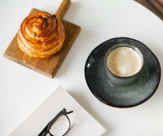 A cup of coffee on the table with a pain au raisin and some glasses