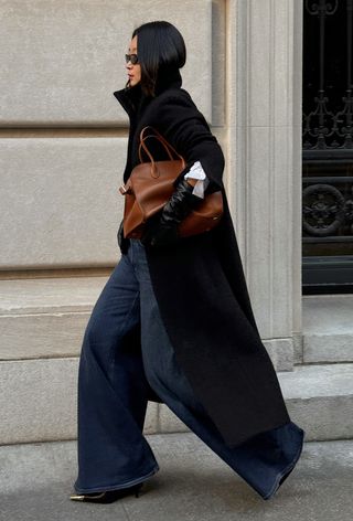 The winter capsule wardrobe item, leather gloves, is shown in a photo of a woman wearing a long black coat over a white button-down shirt styled with leather gloves, a brown bag, wide-leg jeans, and pointed heels