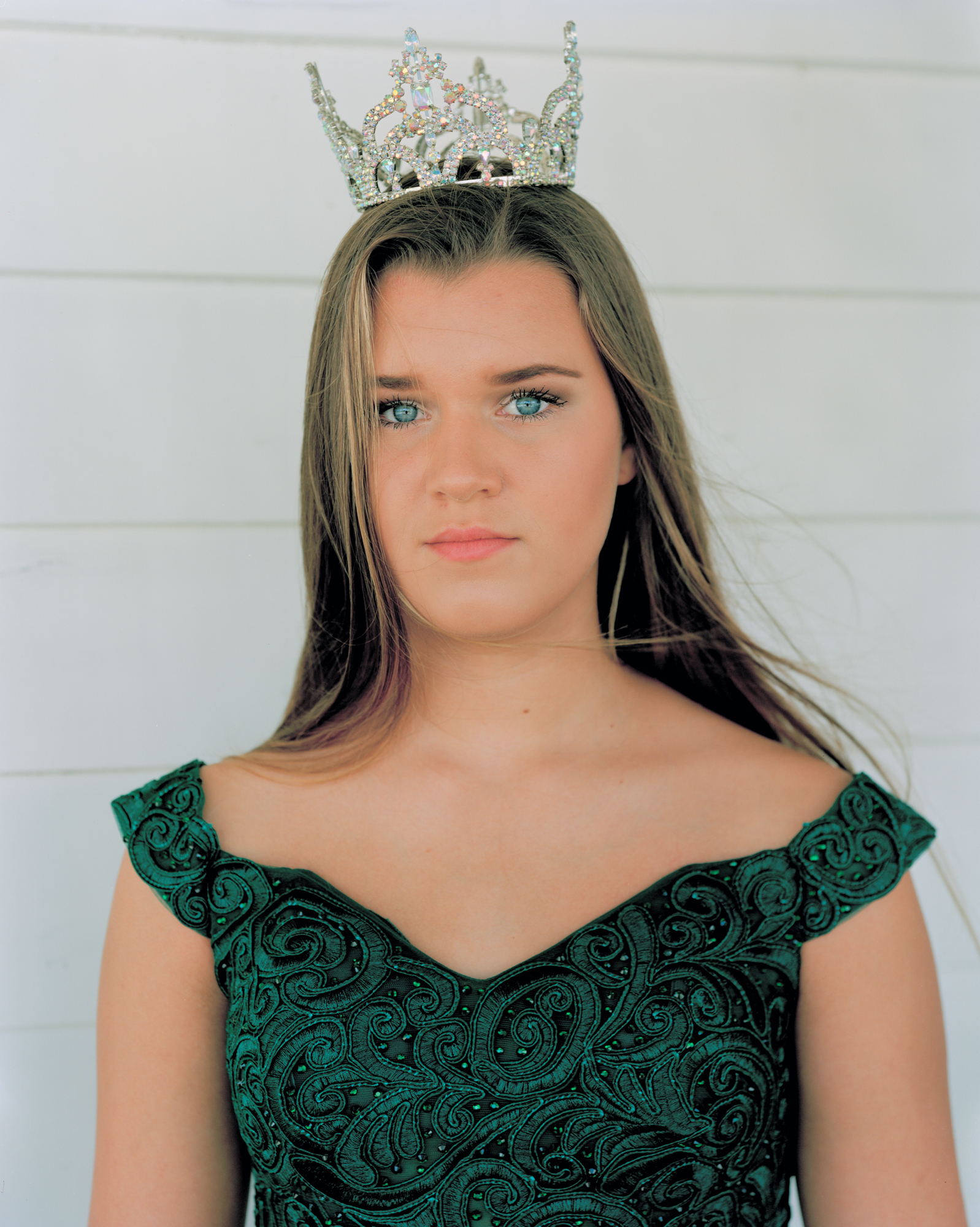 A teenage girl with long hair wears a sparkling tiara and an embroidered green dress against a white wooden wall