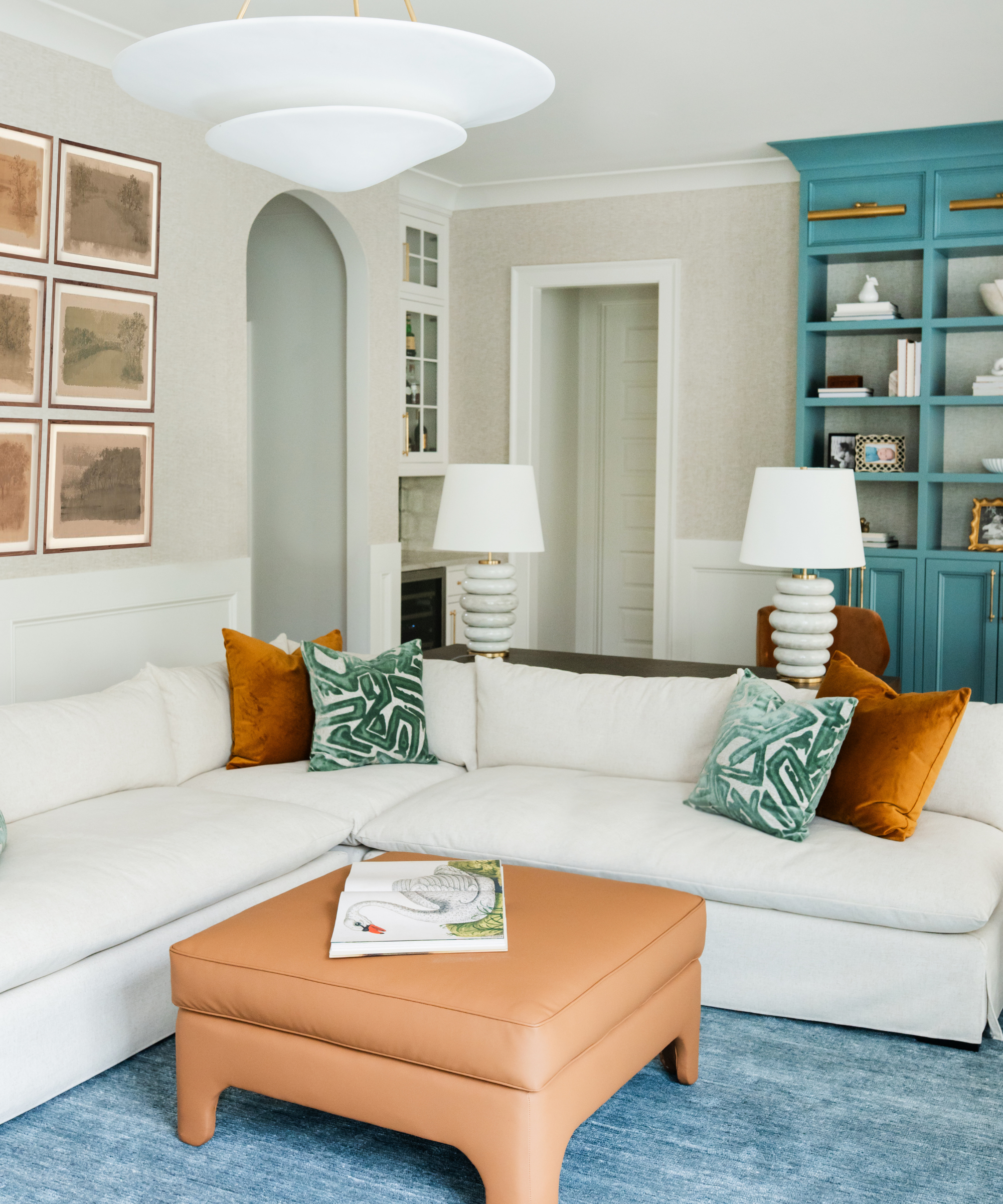 a living room with a white sectional, symmetrical pillows, a tan ottoman, symmetrical lamps, and a teal bookcase in the back