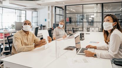 Several mask-wearing office workers sitting at a desk, separated by glass partitions