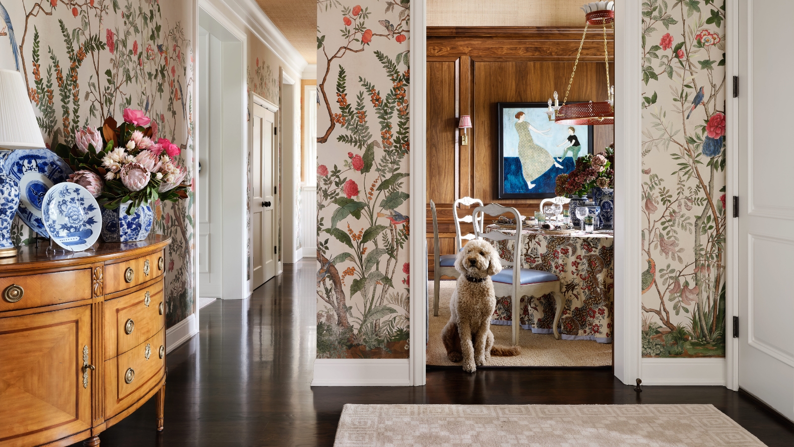 Floral-wallpapered entry with a blue ceiling, antique console, blue-and-white ceramics and a dog framed in the dining-room doorway.