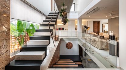 Staircase trends. Large open-plan entryway and living room, chunky marble staircase in black and white, smaller wooden staircase going to lower level, glass panels, statement lighting
