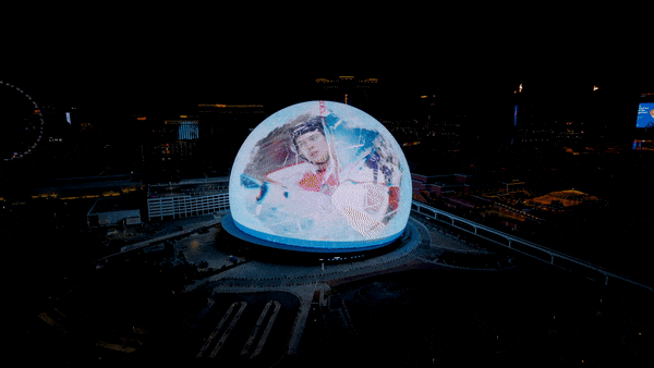 A GIF of a hockey player slapshotting a puck with the NHL Draft logo on it on the exterior of the Sphere. 