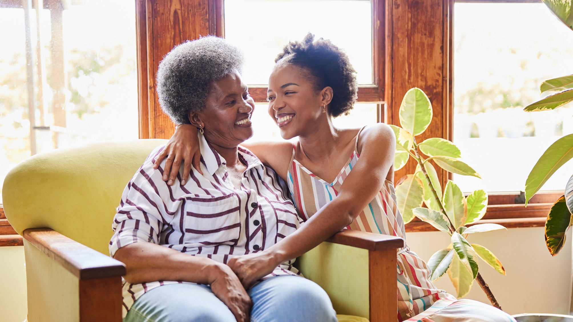 A seated older woman hugs her adult daughter in the living room.