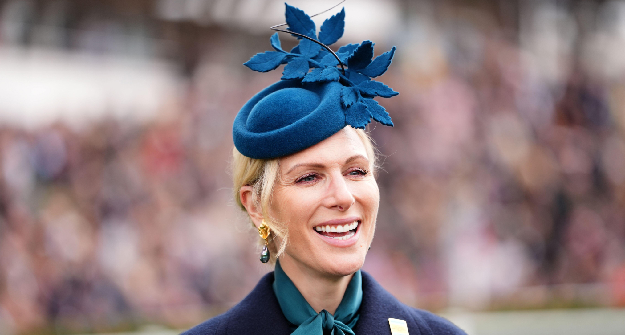 Zara Tindall smiling in a blue hat and tie neck blouse